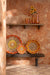 Bruin houten bankje van 190x38x46 cm met decoratieve gevlochten wandmanden tegen een rustieke oranje muur.