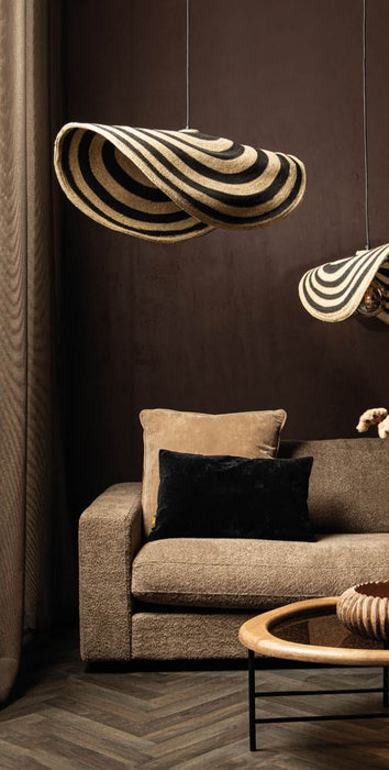 PTMD Loulou hanglamp van rotan met golvend design in beige en zwart boven een bruine bank.