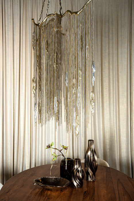 Grote messingkleurige PTMD Wilco hanglamp van 85 cm met decoratieve metalen kettingen boven een houten tafel.