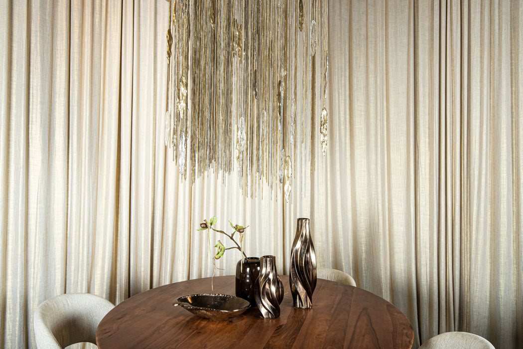 Grote messingkleurige PTMD Wilco hanglamp van gegoten aluminium boven een ronde houten tafel met decoratieve vazen.